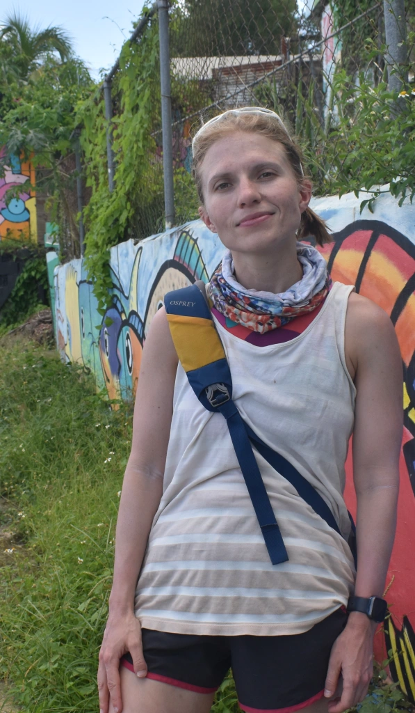 a white woman in a tan and white striped tank top and gym shorts leans against a wall decorated with a colorful mural, in an alleyway overgrown with gracc.